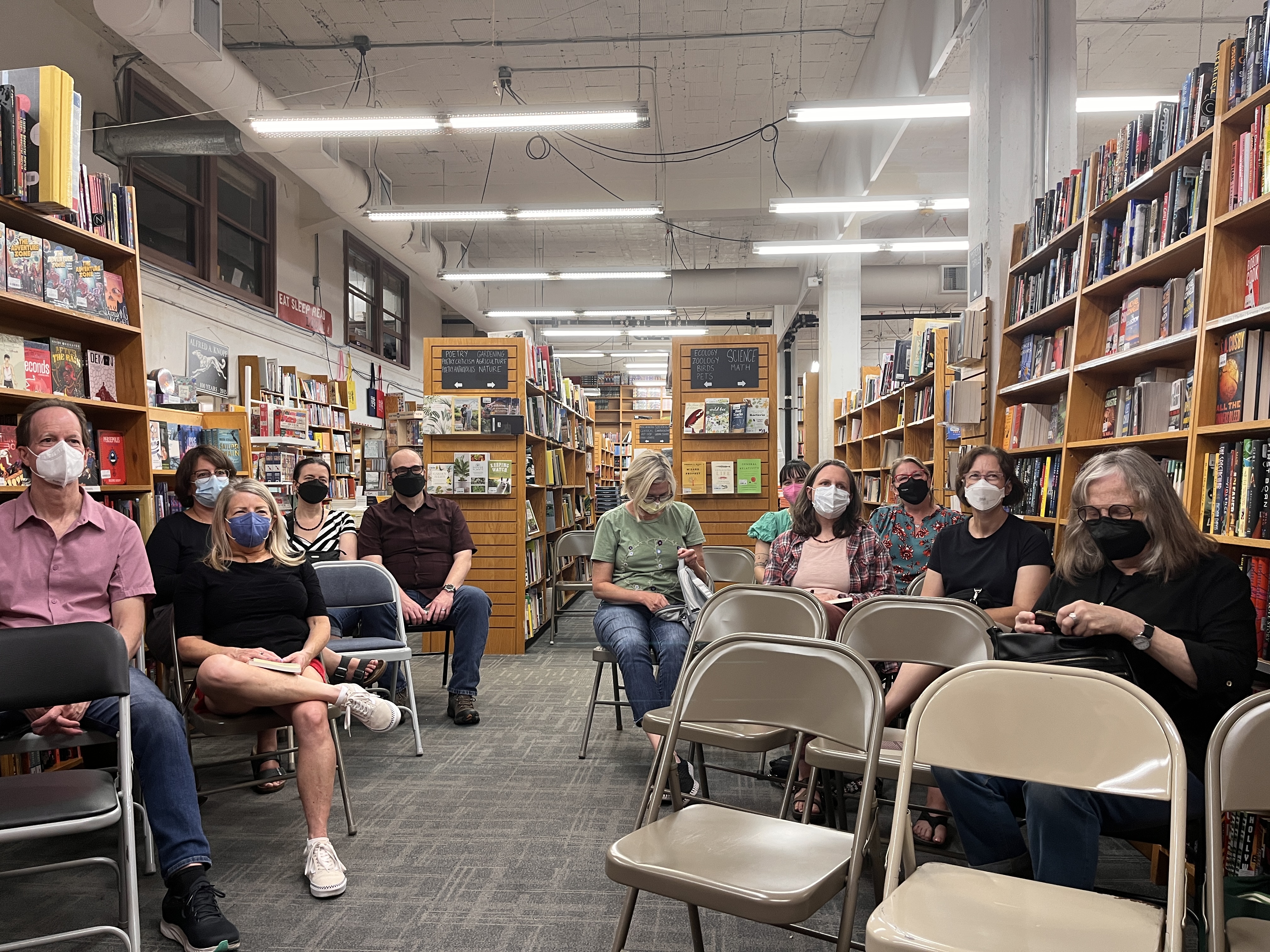 Audience of people seated on folding chairs wearing masks and surrounded by bookshelves.