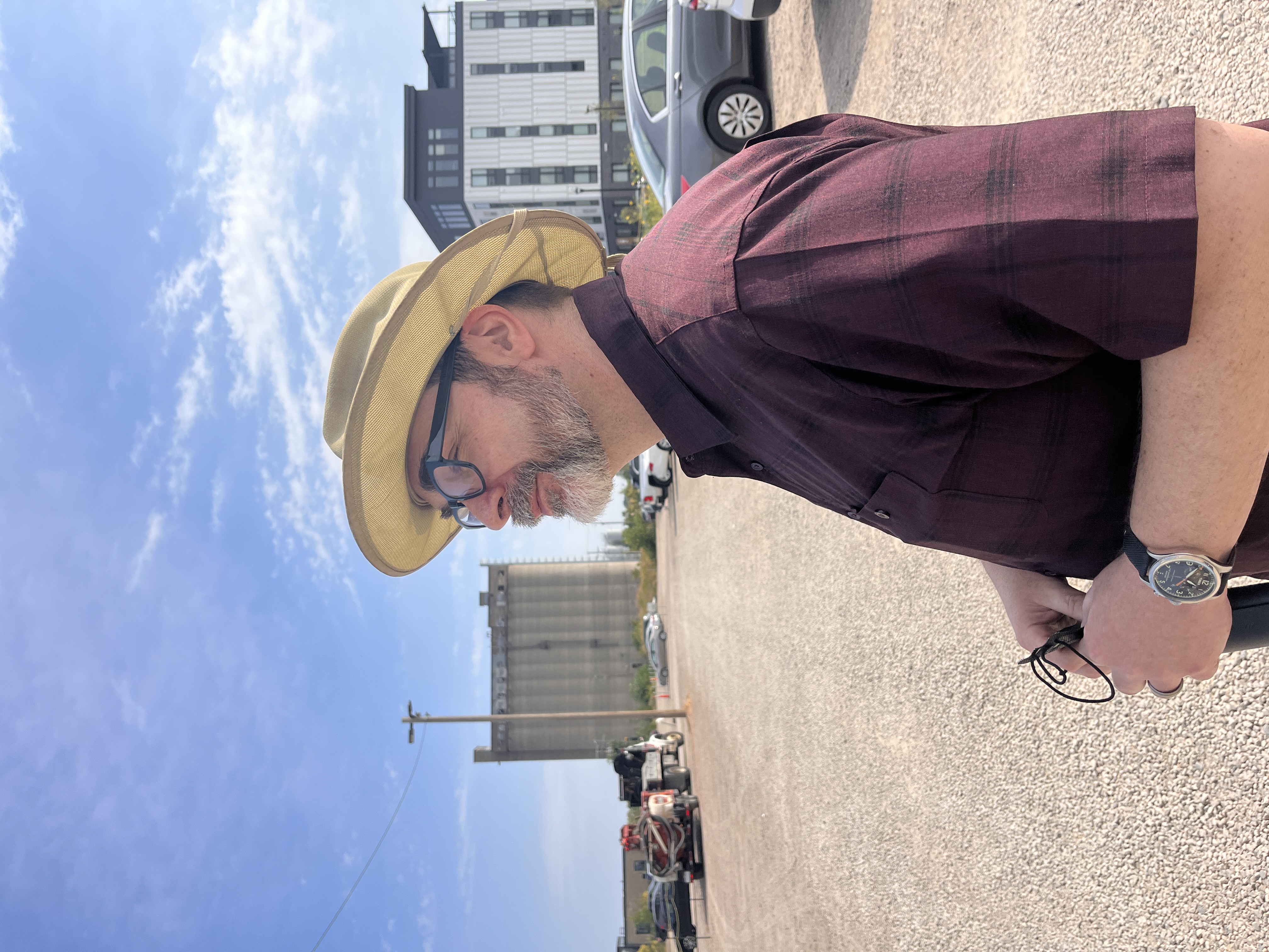 A white man wearing a brimmed hat and glasses, staring to the left with a concrete grain storage building and huge blue sky behind him.