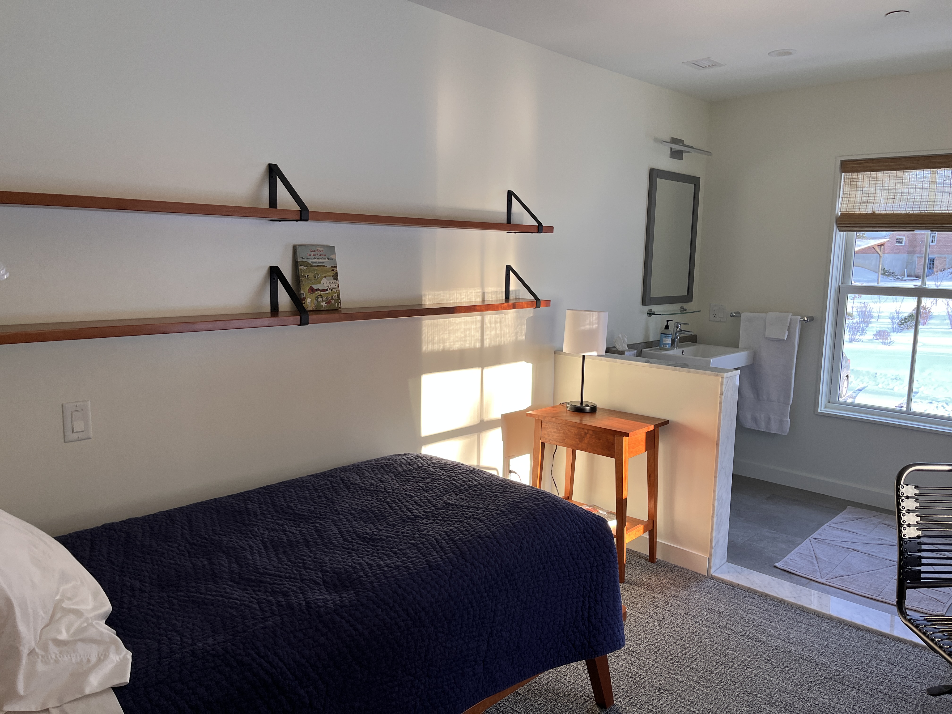 A room lit by natural like with wooded shelves, a nightstand, and a bed with a blue bedspread with a bathroom nook beyond.