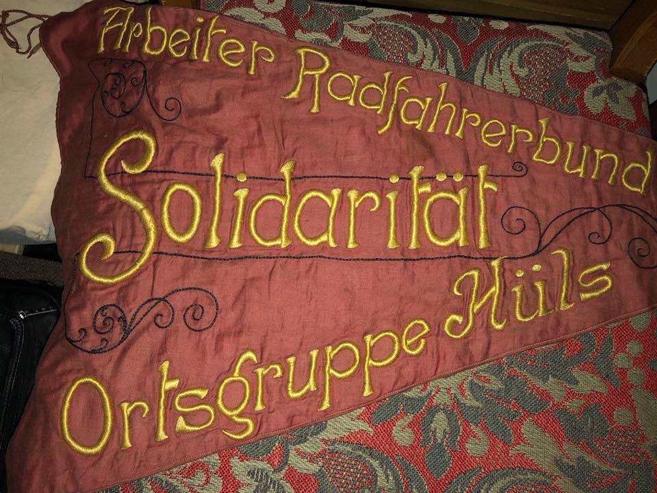 Red flag with embroidererd gold letters Socialist Workers’ Bicycle Club. It says “Solidarity” in the middle and the village chapter of Hüls, their neighborhood