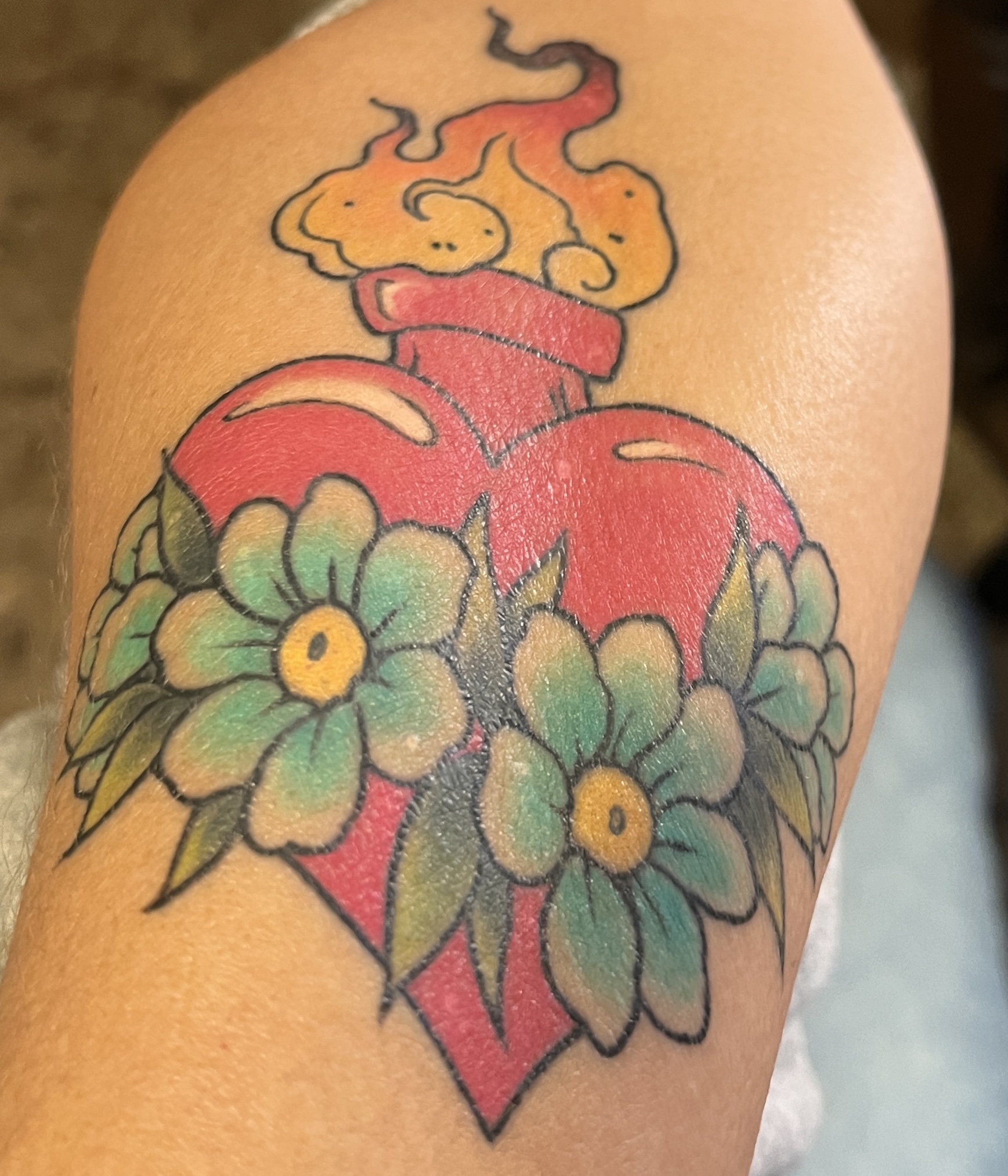 A tattoo on a tan-colored forearm of a red heart crossed with florals and with flame coming out of the top.