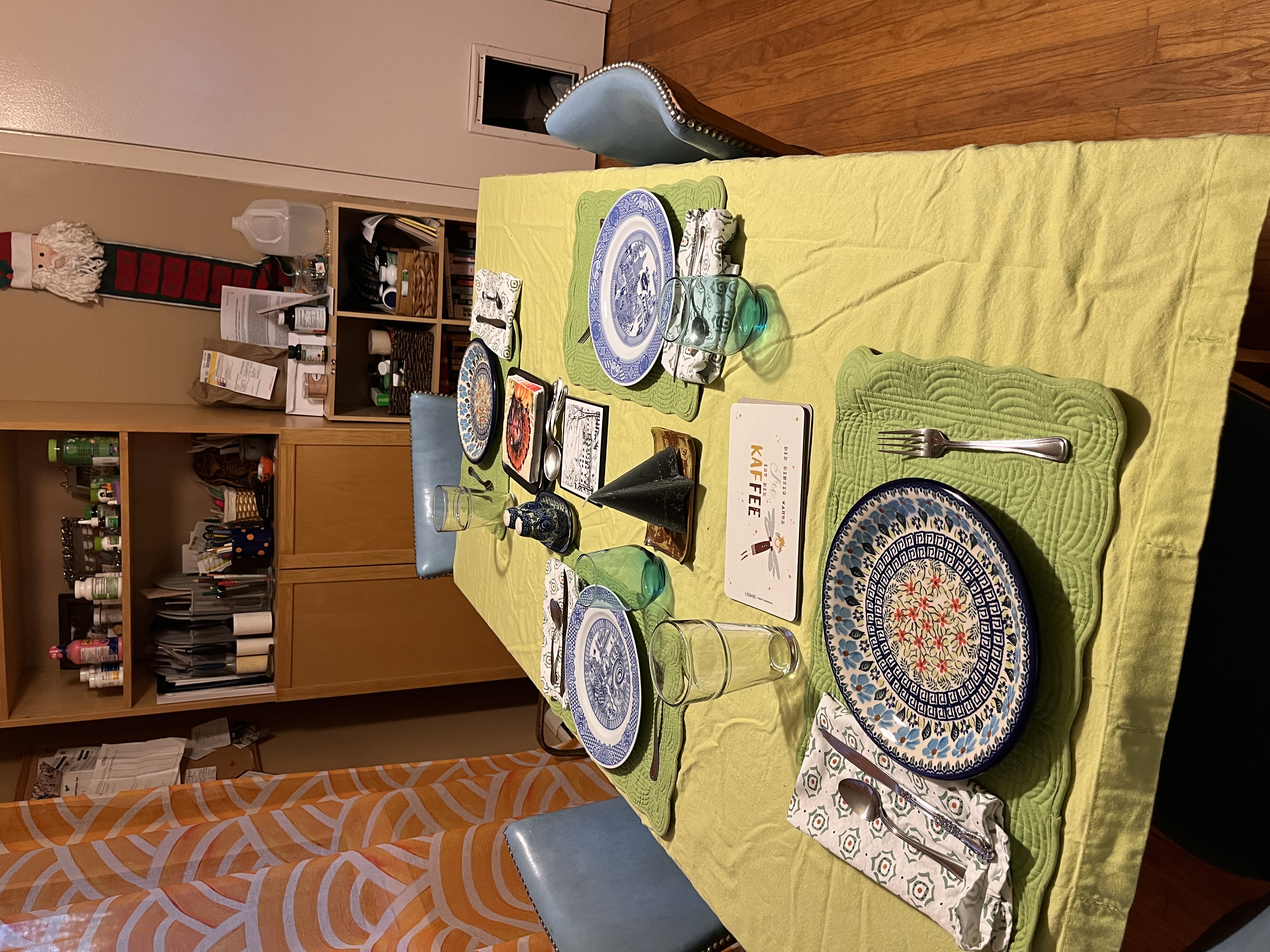 A table set with a mix of blue and white plates on a lime-green cloth, sorta wrinkly, on green placemats, four chairs, with a shelf behind it cluttered with pill bottles and papers, and a curtain hanging to the left that is gauzy orange.