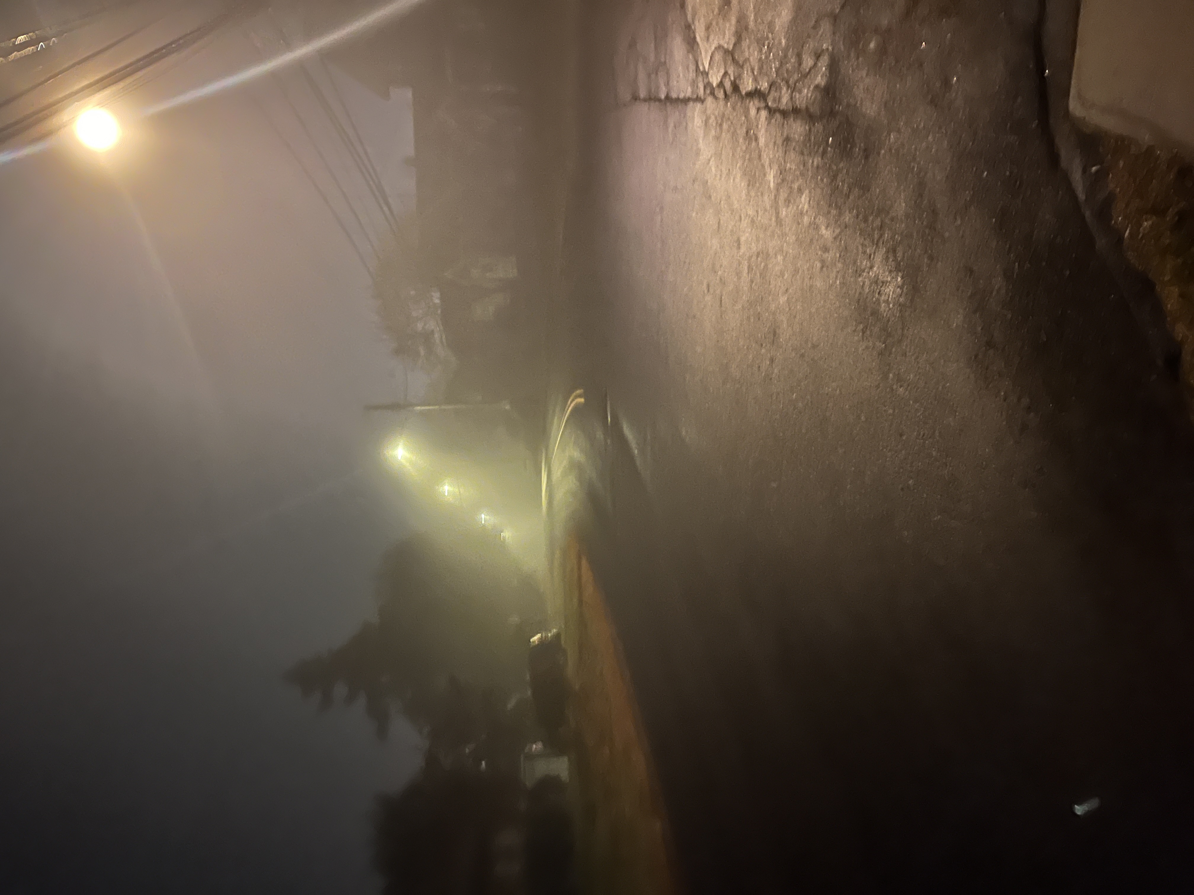 A roadway curving to the left, with powerlines above, in the darkness, and the streetlights in fog trace a trail.