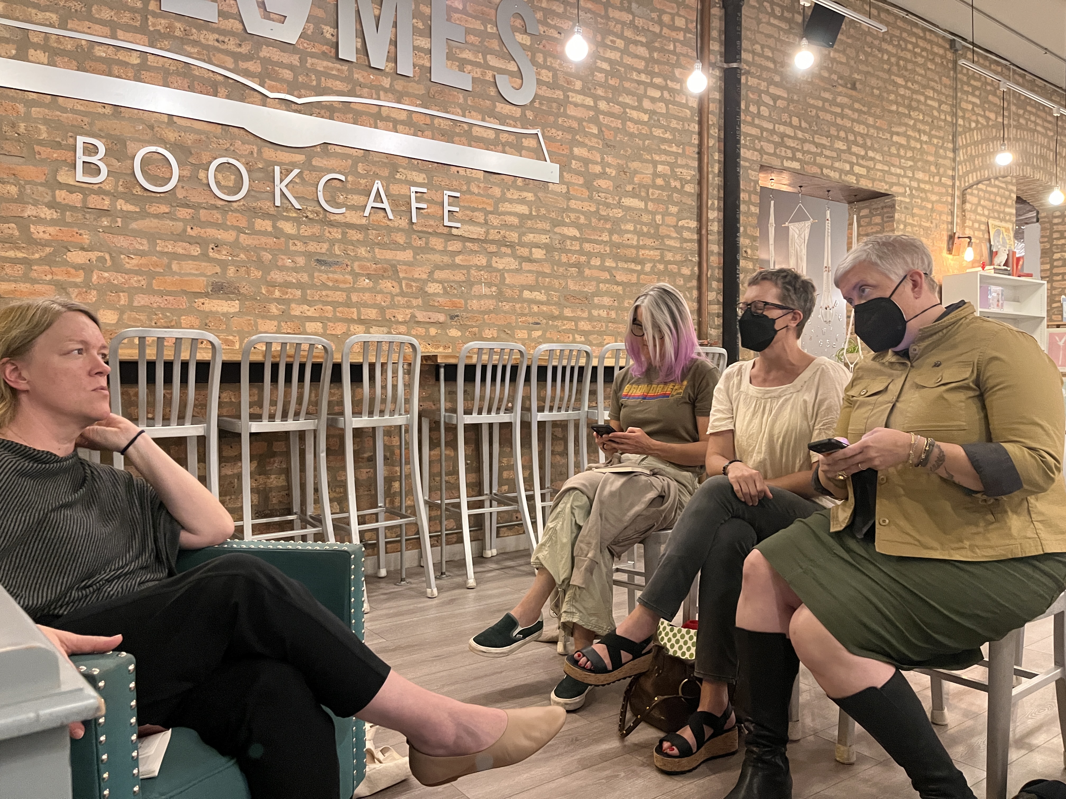 Megan, Mary, Martha and Eirin sitting in front of a long brick wall with Volumes Bookcafe in metal letters with a row of chairs near a counter.