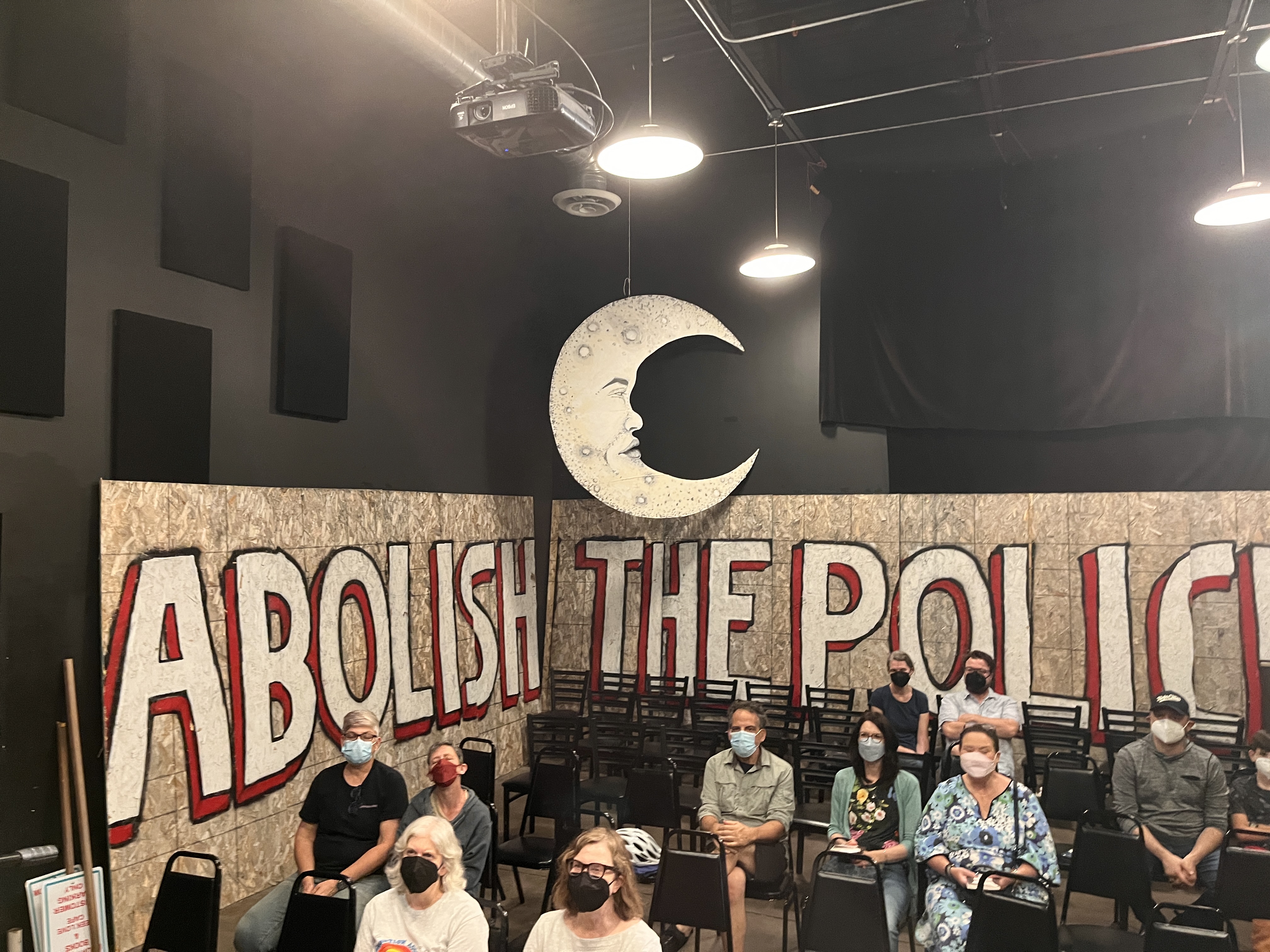 A shot from stage down into the audience in a blackbox theater, with ten people wearing masks in chairs, and the slogan Abolish the Police on the wall behind with a moon hanging down.