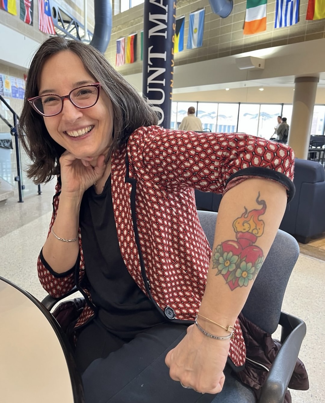 Pic taken by Jim Reese at Mount Marty University in Yankton, SD, where I visited at the end of last week.​ I am sitting in a cafeteria, smiling and wearing a red patterned blazer, with one arm propped up, fist on my leg, showing a sacred heart tattoo that looks a bit like a molotov cocktail on my forearm. 