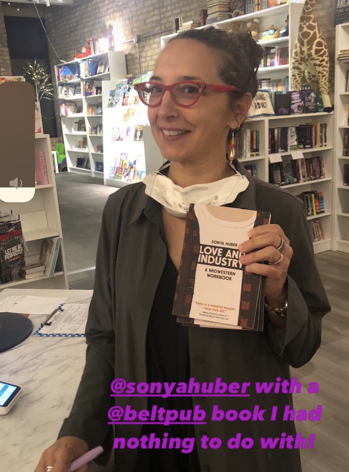 Sonya with red glasses holding copy of Love and Industry with white shelves filled with books in the background along a brick wall.