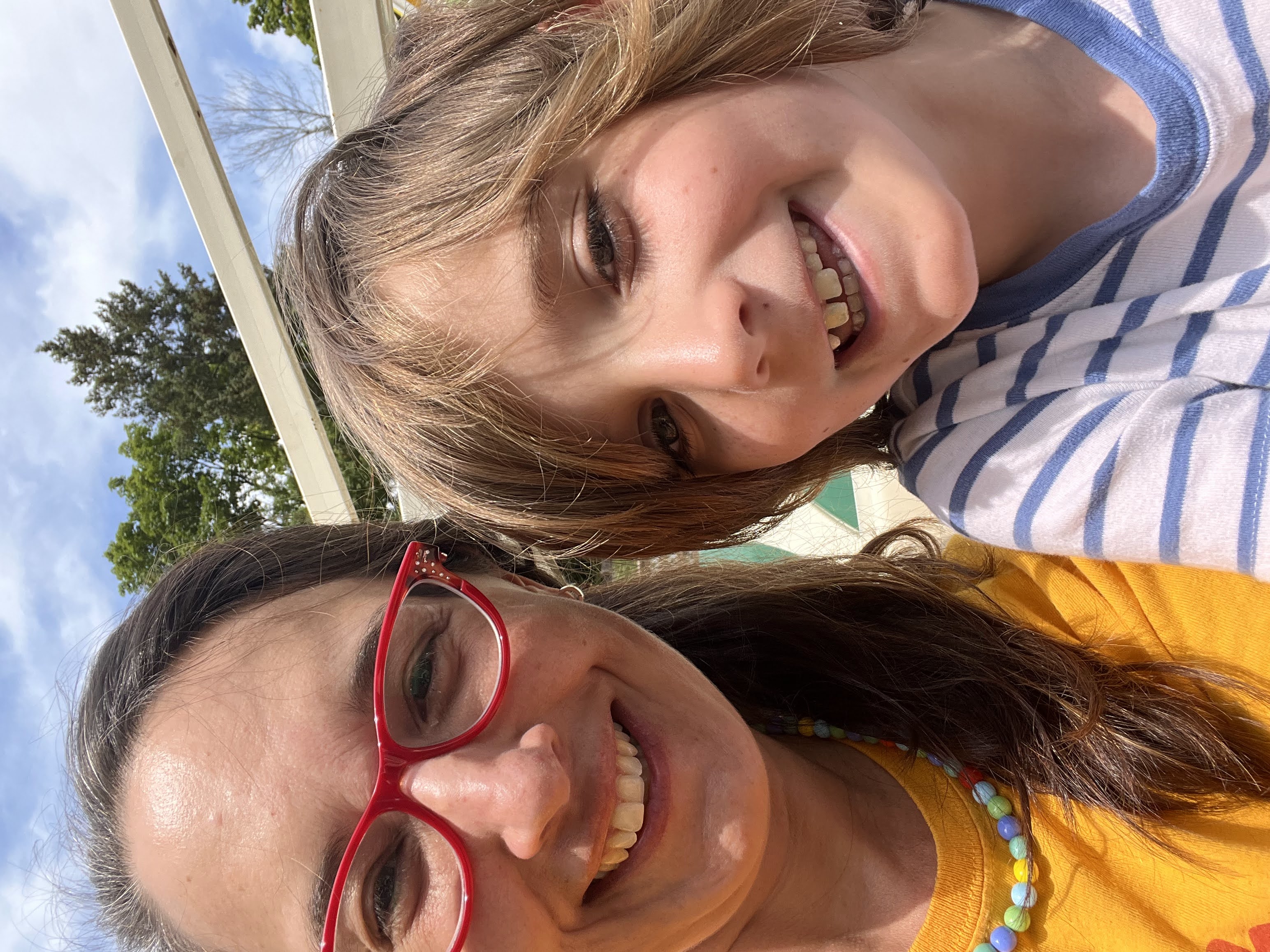 Sonya with long dark hair with gray in red glasses and a colorful shirt and necklace, next to Nathaniel who has longish hair and an impish grin, outside with trees in the background