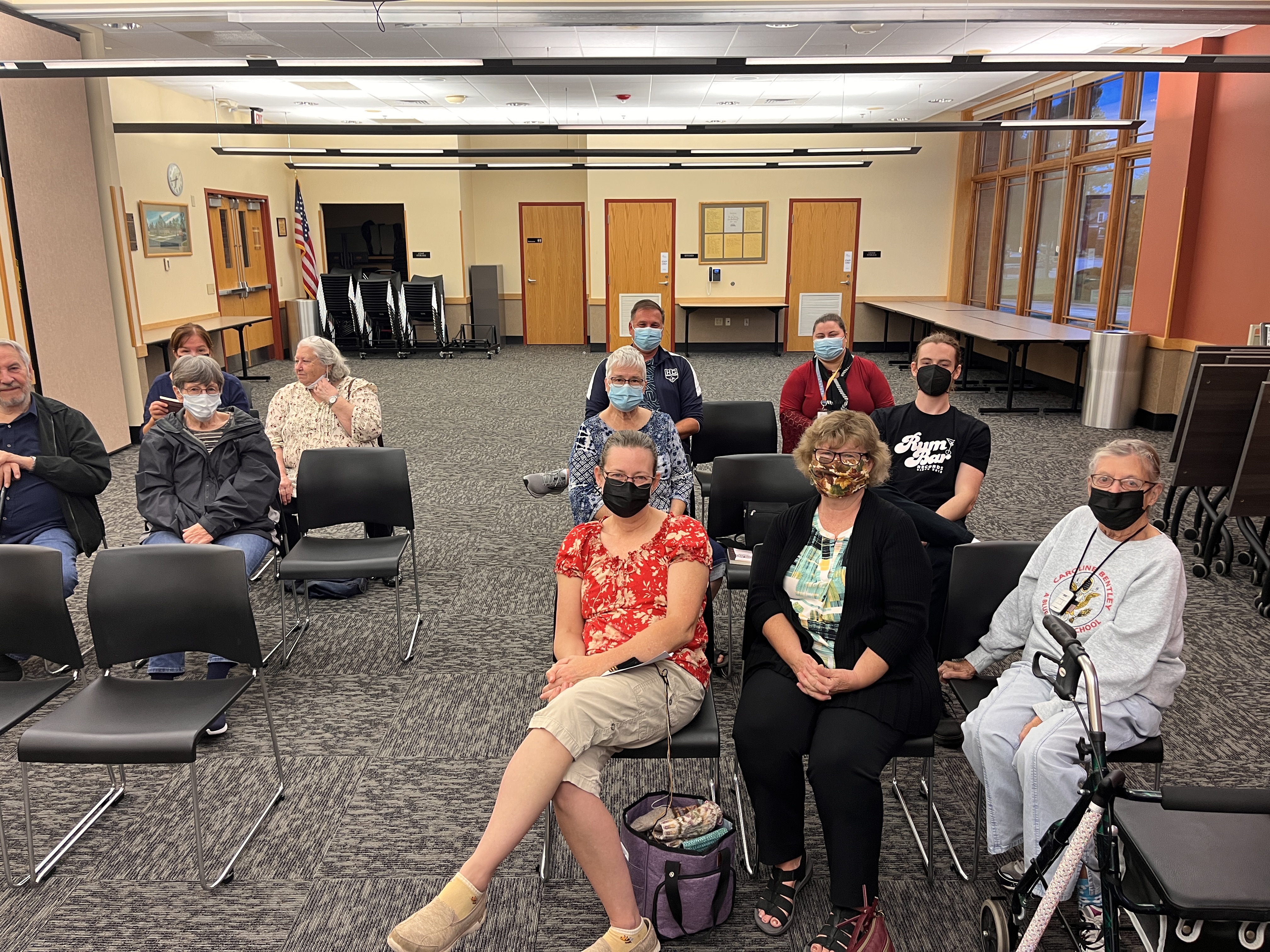 Function room with three rows of seats in two sections, with masked folks sitting in the seats and fluorescent lights overhead